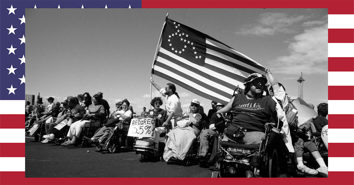 Disability Rights protestors in a line, one holding the likeness of an American Flag with the stars in the shape of a person in a wheelchair