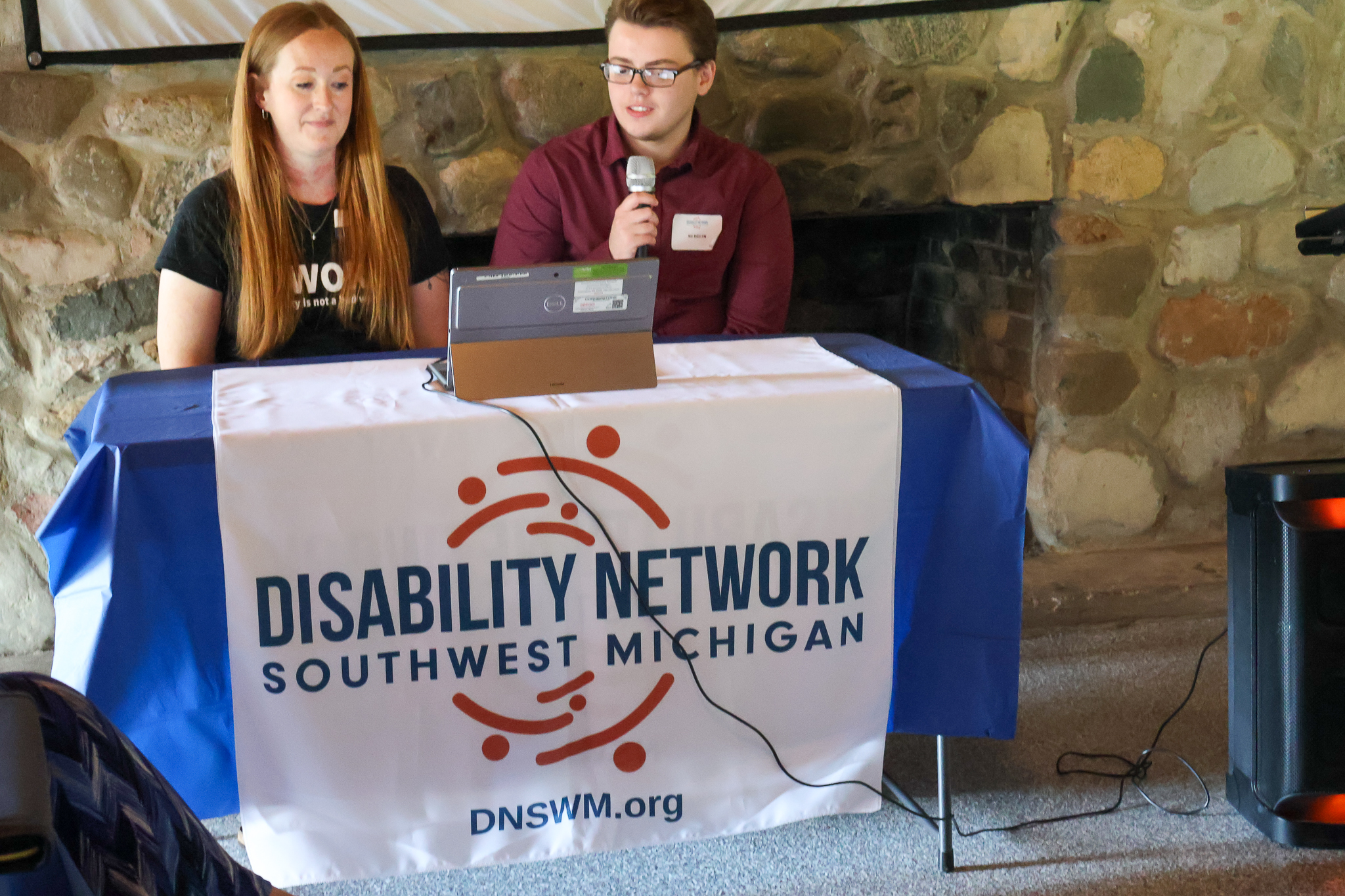 A white woman and a white man seated at a table with microphones with a Disability Network table runner.