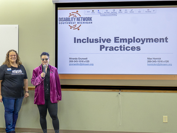 A white woman and a white non-binary person standing in front of a projector screen where a Disability Network presentation on Inclusive Employment Practices is displayed.