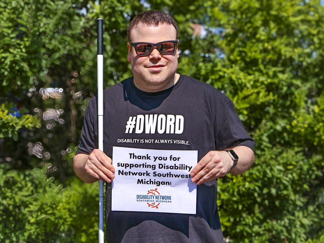 A blind man wearing a black #DWord shirt and holding a white cane plus a sign that says Thank you for supporting Disability Network Southwest Michigan.