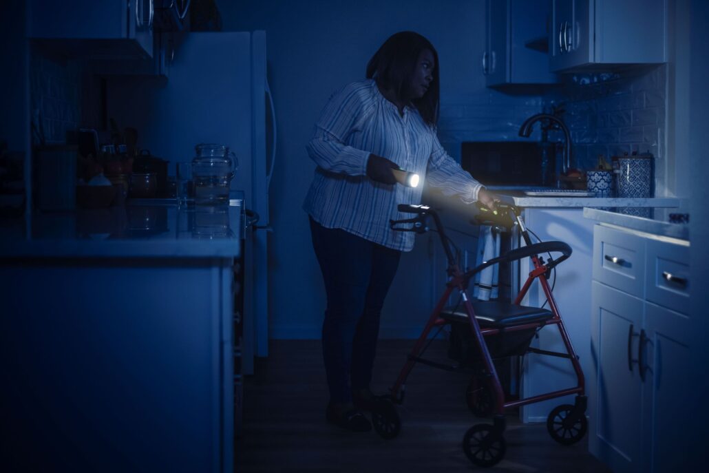 Woman using a walker, standing in a dark kitchen with a flashlight