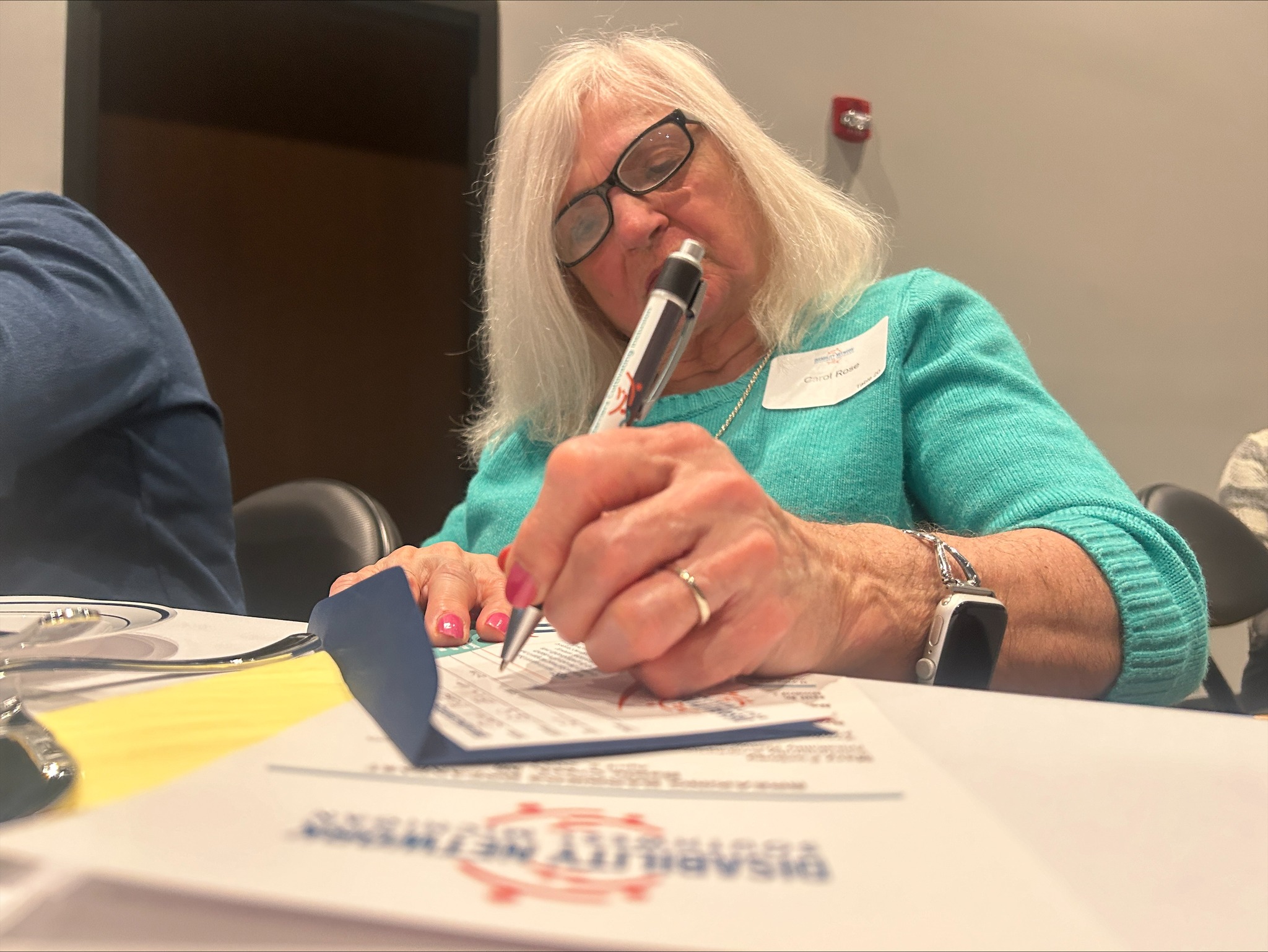 An older woman with white hair writes on a pledge card.