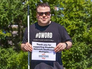 A blind man wearing a black #DWord shirt and holding a white cane plus a sign that says Thank you for supporting Disability Network Southwest Michigan.