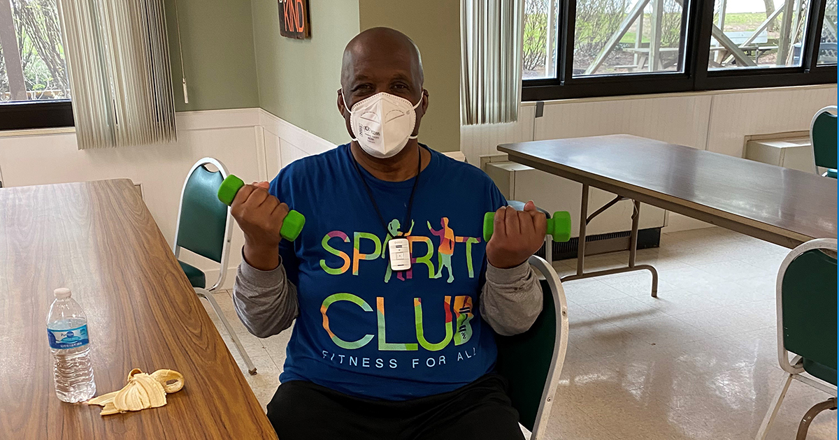A man seated, wearing mask and living dumb bells, his shirt reads "Spirit Club"